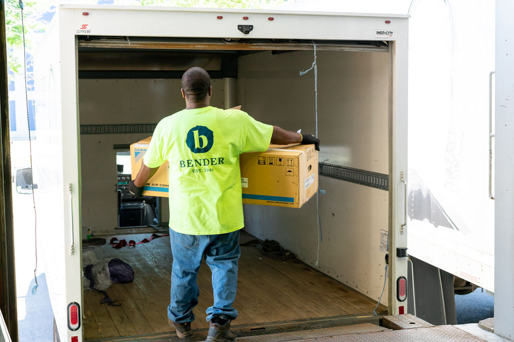 A person wearing a Bender t-shirt carrying a large Daikin box into a box truck, work boots, work gloves